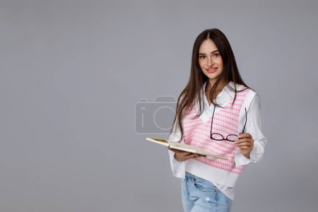 Téléchargez les photos : Femme brune heureuse avec des lunettes et livre souriant sur fond gris. espace de copie - en image libre de droit