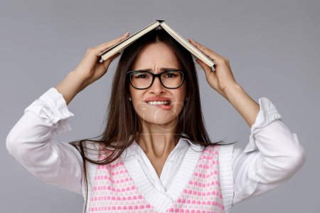 Photo for Puzzled brunette woman in glasses feels sorry biting lips on grey background. oops. girl with book on head - Royalty Free Image