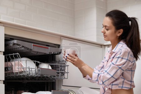 Foto de Mujer atractiva taza de descarga de la máquina de lavavajillas integrada automática abierta con utensilios limpios en el interior en la cocina blanca moderna. - Imagen libre de derechos