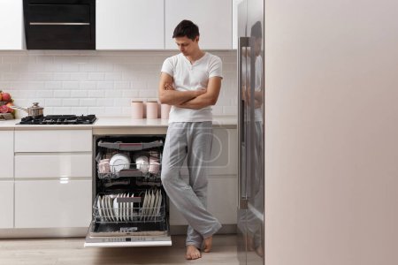 Foto de Marido guapo hombre usando lavavajillas y lavando platos en la cocina moderna blanca. espacio de copia - Imagen libre de derechos