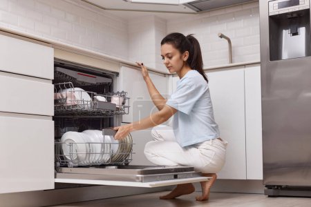 Téléchargez les photos : Jolie femme chargement dished pour ouvrir automatique lave-vaisselle intégré machine avec ustensiles propres à l'intérieur dans la cuisine blanche moderne. - en image libre de droit