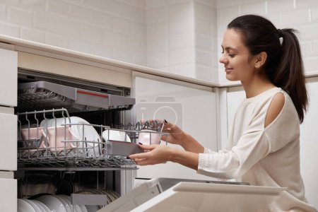 Photo for Attractive woman unloading cup from open automatic built-in dishwasher machine with clean utensils inside in modern white kitchen. - Royalty Free Image