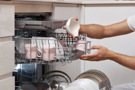 Téléchargez les photos : Plaques de déchargement à la main mâles de la machine à laver le vaisselle automatique ouverte avec des ustensiles propres à l'intérieur dans la cuisine. - en image libre de droit