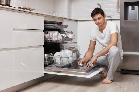 Foto de Hombre guapo poner la tableta detergente en el lavavajillas en la cocina - Imagen libre de derechos