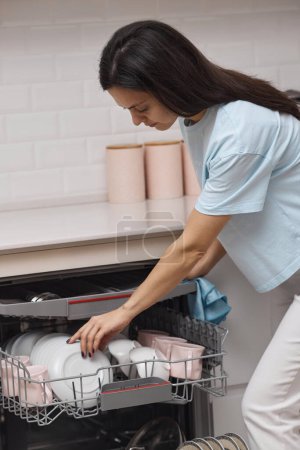 Photo for Pretty woman unloading from open automatic built-in dishwasher machine with clean utensils inside in modern white kitchen. - Royalty Free Image