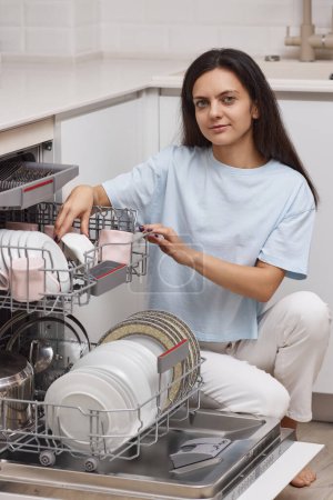 Foto de Mujer bonita descarga de la máquina de lavavajillas incorporado automático abierto con utensilios limpios en el interior en la cocina blanca moderna. - Imagen libre de derechos
