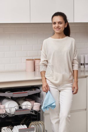 Photo for Young smiling housewife woman holding blue towel and using modern dishwasher for wash dishes at white modern kitchen. - Royalty Free Image