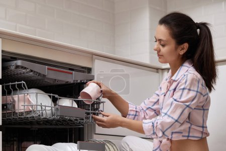 Téléchargez les photos : Attrayant femme déchargement tasse de lave-vaisselle automatique ouvert avec ustensiles propres à l'intérieur dans la cuisine blanche moderne. - en image libre de droit