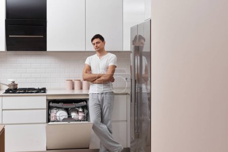 Photo for Handsome guy using dishwasher and washing dishes in white modern kitchen. copy space - Royalty Free Image
