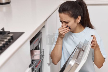 Téléchargez les photos : Femme sent odeur étrange et terrible provenant de la machine à lave-vaisselle dans la cuisine - en image libre de droit