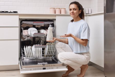 Foto de Mujer vierte ayuda de enjuague en el compartimiento del lavavajillas en la cocina blanca moderna - Imagen libre de derechos