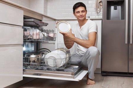 Foto de Apuesto hombre positivo saca platos limpios del lavavajillas en la cocina moderna blanca - Imagen libre de derechos