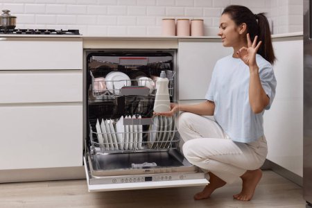 Foto de Mujer vierte ayuda de enjuague en el compartimiento del lavavajillas en la cocina blanca moderna - Imagen libre de derechos