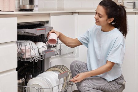 Téléchargez les photos : Attrayant femme déchargement tasse de lave-vaisselle automatique ouvert avec ustensiles propres à l'intérieur dans la cuisine blanche moderne. - en image libre de droit