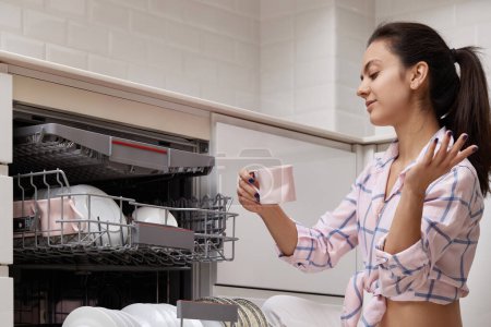 Foto de Mujer atractiva taza de descarga de la máquina de lavavajillas integrada automática abierta con utensilios limpios en el interior en la cocina blanca moderna. - Imagen libre de derechos