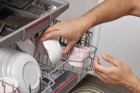Foto de Placas de descarga de mano masculina de la máquina lavavajillas automática abierta con utensilios limpios en el interior de la cocina. - Imagen libre de derechos