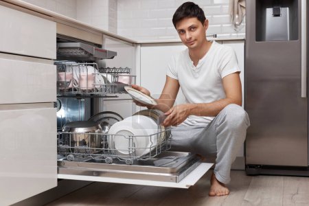 Foto de Apuesto hombre positivo saca platos limpios del lavavajillas en la cocina moderna blanca - Imagen libre de derechos