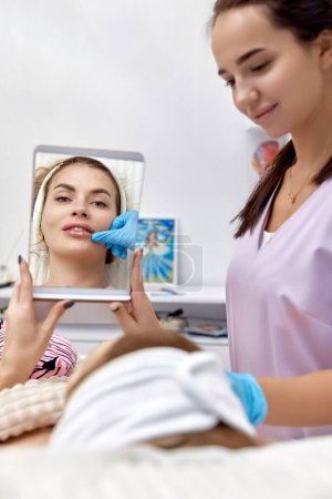 Téléchargez les photos : Belle femme se regarde dans le miroir après l'augmentation des lèvres procédure dans la clinique médicale de beauté. - en image libre de droit