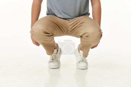 Téléchargez les photos : Homme portant des baskets blanches et un pantalon beige décontracté accroupi sur fond de studio. vue de face - en image libre de droit