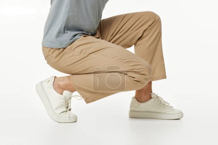 Téléchargez les photos : Homme portant des baskets blanches et un pantalon beige décontracté accroupi sur fond de studio. vue latérale - en image libre de droit