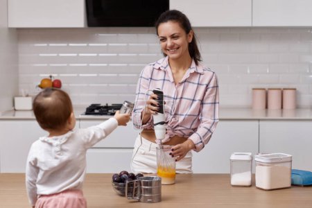 Téléchargez les photos : Mère souriante avec sa petite fille préparant une tarte aux prunes à la cuisine blanche. cuisiner ensemble - en image libre de droit