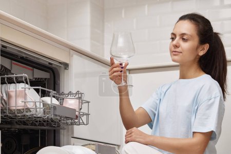 Foto de Mujer bonita toma copas de vino del lavavajillas y sonríe en la cocina moderna. - Imagen libre de derechos