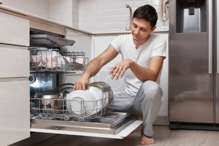 Téléchargez les photos : Bel homme positif mettre des assiettes dans la machine à lave-vaisselle dans la cuisine - en image libre de droit