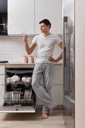 Foto de Hombre feliz sacando vidrio limpio de la máquina lavavajillas en la cocina - Imagen libre de derechos