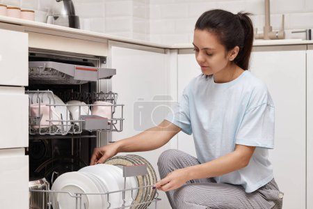 Téléchargez les photos : Femme heureuse déchargeant la plaque de lave-vaisselle automatique ouverte avec des ustensiles propres à l'intérieur dans la cuisine blanche moderne. - en image libre de droit