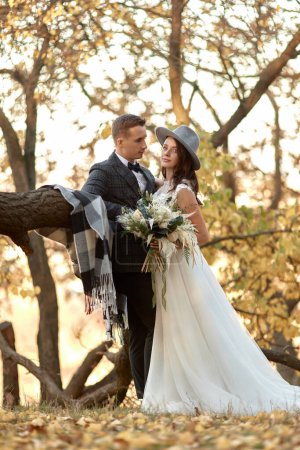 Téléchargez les photos : Belle mariée élégante heureuse en chapeau et marié debout en plein air en automne - en image libre de droit