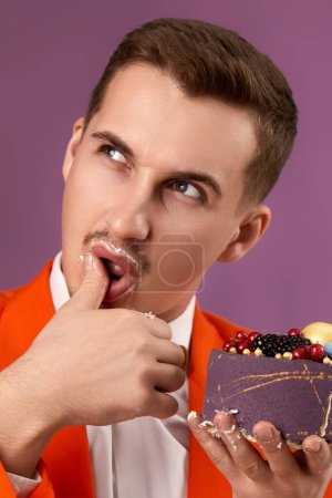 Foto de Cumpleañero con chaqueta naranja sosteniendo pastel mirando a la cámara siendo encantadora y sexy sobre fondo púrpura - Imagen libre de derechos