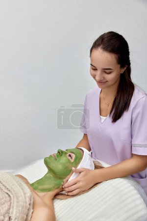 Foto de Esteticista femenina aplica la máscara a la cara de la hermosa mujer en el salón de spa. cuidado facial de piel - Imagen libre de derechos
