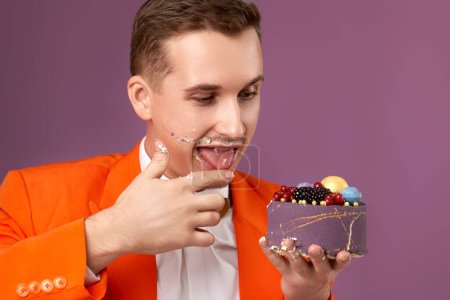 Photo for Birthday man in orange jacket eating yummy cake on purple background - Royalty Free Image