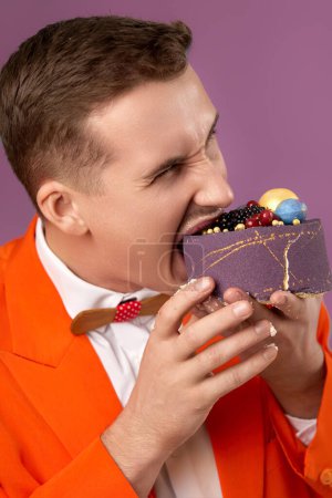 Téléchargez les photos : Bel homme anniversaire en veste orange mord gâteau sur fond violet - en image libre de droit