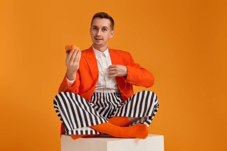 Foto de Extravagante joven divertido en chaqueta naranja y pantalones a rayas con cupcake sobre fondo naranja. - Imagen libre de derechos