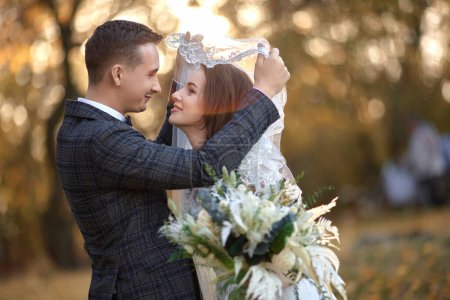 Téléchargez les photos : Heureux mariés profitant de moments romantiques sous le voile et se regardant sur fond naturel dans la journée ensoleillée - en image libre de droit