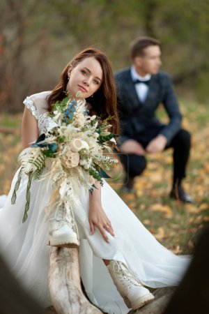 Photo for Beautiful bride in white boots sitting on tree and holding autumn bouquet. blur groom on the background - Royalty Free Image