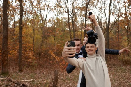 Téléchargez les photos : Photographe de mariage avec mariée et marié prendre selfie avec téléphone portable dans la nature - en image libre de droit