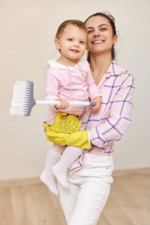 Photo for Happy mother housewife is holding cute baby girl and doing housework at home - Royalty Free Image