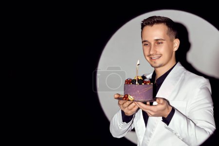 Téléchargez les photos : Élégant homme caucasien en costume blanc smoking tenant gâteau avec bougie dans le cercle de lumière, espace de copie. - en image libre de droit