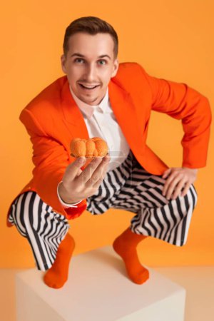 Téléchargez les photos : Jeune homme drôle extravagant en veste orange et pantalon rayé tenant cupcake sur fond orange. - en image libre de droit