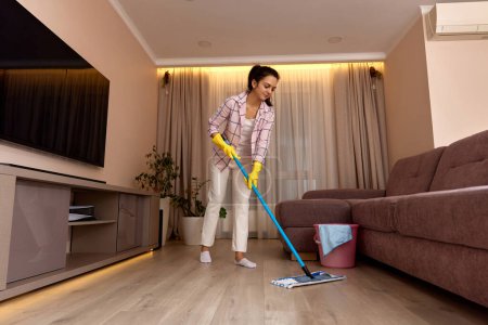 Téléchargez les photos : Nettoyage et nettoyage du sol de la jeune femme au salon le soir, ménage quotidien - en image libre de droit