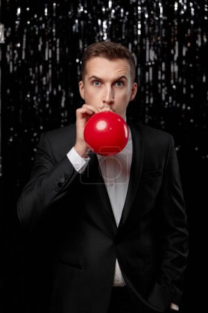Photo for Caucasian man in black jacket blowing red air balloon on glitter background. new year party - Royalty Free Image