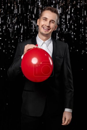 Téléchargez les photos : Homme souriant en veste noire tenant ballon à air rouge sur fond pailleté foncé. nouvelle année fête - en image libre de droit