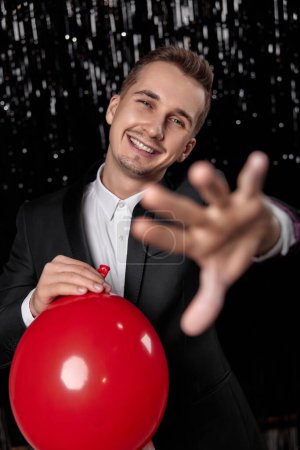 Foto de Hombre positivo en chaqueta negra con globo de aire rojo sobre fondo de brillo oscuro. fiesta de año nuevo - Imagen libre de derechos