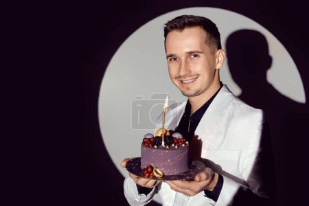 Téléchargez les photos : Élégant homme souriant en costume blanc smoking tenant gâteau avec bougie dans le cercle de lumière, espace de copie. Fête - en image libre de droit