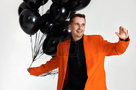 Téléchargez les photos : Gars souriant en veste orange avec des ballons à air noir sur fond blanc. fête d'anniversaire - en image libre de droit