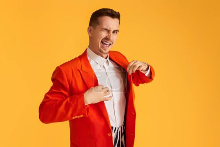 Photo for Stylish winking young man in orange jacket and striped pants on vibrant yellow background. - Royalty Free Image