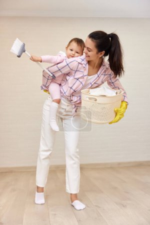Foto de Feliz madre ama de casa está sosteniendo lindo bebé niña y cesta con lavandería en casa, familia feliz - Imagen libre de derechos