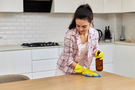 Foto de Mujer joven en guantes limpiando y limpiando mesa de madera con paño de microfibra. tareas en casa. Servicio de limpieza - Imagen libre de derechos